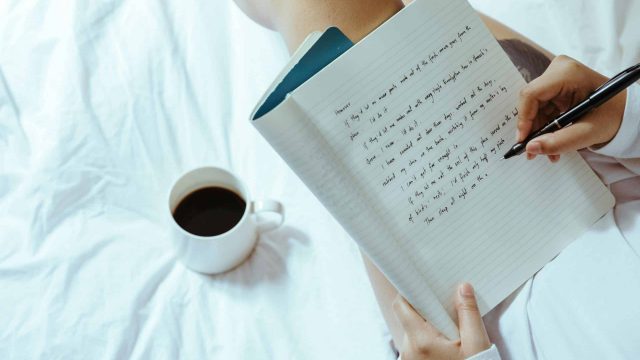 Top view of unrecognizable woman sitting on bed with legs near cup of coffee and writing on notepad with pen while resting at home